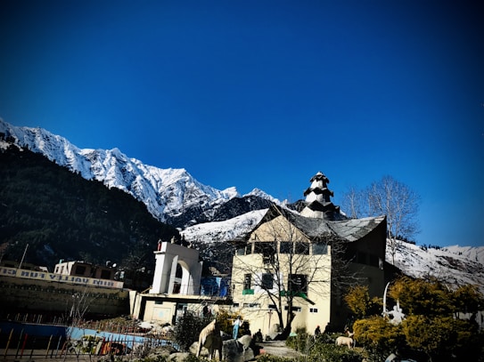 A scenic mountainscape features a striking clear blue sky above snow-capped peaks, creating a dramatic backdrop. In the foreground, a distinctively designed building, possibly a temple or cultural structure, is visible with its intricate architecture. Bare trees and green bushes surround the structure, adding to the picturesque setting.