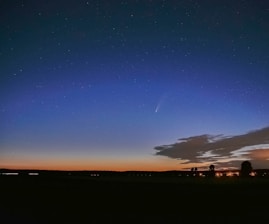A serene night sky features a brilliant comet with a visible tail streaking across it. The sky transitions from a deep blue at the top to a soft orange along the horizon. Below the sky, silhouettes of trees and distant lights from buildings provide a tranquil foreground. Scattered clouds add texture and depth to the scene.
