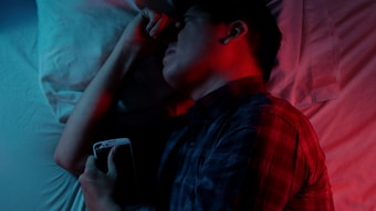 A person lies in bed holding a smartphone, with a red and blue light casting contrasting shadows. The person appears to be asleep or deep in thought. A white pillow and sheet are visible in the background.