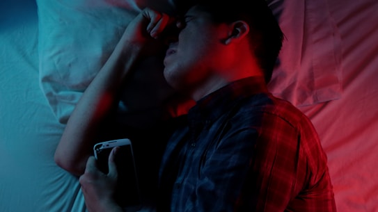 A person lies in bed holding a smartphone, with a red and blue light casting contrasting shadows. The person appears to be asleep or deep in thought. A white pillow and sheet are visible in the background.