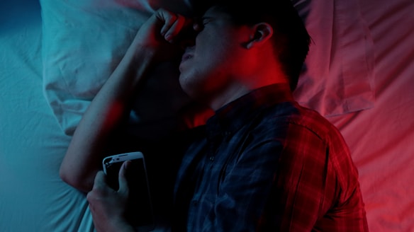 A person lies in bed holding a smartphone, with a red and blue light casting contrasting shadows. The person appears to be asleep or deep in thought. A white pillow and sheet are visible in the background.