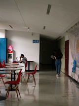 A hallway with tables and chairs, featuring a person sitting at a table using a laptop. The chairs are red and stylish, and there are accessibility signs on the wall. Another person walks towards a dimly lit area holding something in their hand.