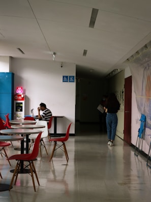 A hallway with tables and chairs, featuring a person sitting at a table using a laptop. The chairs are red and stylish, and there are accessibility signs on the wall. Another person walks towards a dimly lit area holding something in their hand.