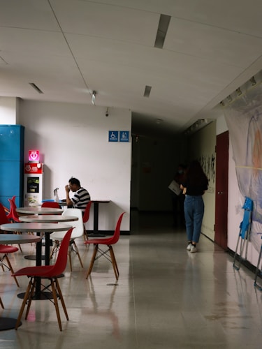 A hallway with tables and chairs, featuring a person sitting at a table using a laptop. The chairs are red and stylish, and there are accessibility signs on the wall. Another person walks towards a dimly lit area holding something in their hand.