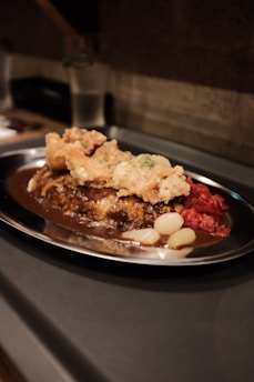 A serving of Japanese curry on a stainless steel plate, topped with fried ingredients and accompanied by pickled vegetables.