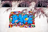 A mural featuring the word 'PACE' in large, bold blue letters surrounded by various national flags. The flags represent different countries, creating a sense of global unity. Branches with pink leaves hang above the mural, adding a touch of nature to the scene.