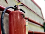 A close-up view of a red fire extinguisher mounted on a wall. Water droplets are visible on its surface, suggesting it was recently used or exposed to moisture. The metal handle and pressure gauge are also visible. The background includes red pipes and a concrete wall.