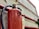 A close-up view of a red fire extinguisher mounted on a wall. Water droplets are visible on its surface, suggesting it was recently used or exposed to moisture. The metal handle and pressure gauge are also visible. The background includes red pipes and a concrete wall.
