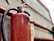 A close-up view of a red fire extinguisher mounted on a wall. Water droplets are visible on its surface, suggesting it was recently used or exposed to moisture. The metal handle and pressure gauge are also visible. The background includes red pipes and a concrete wall.