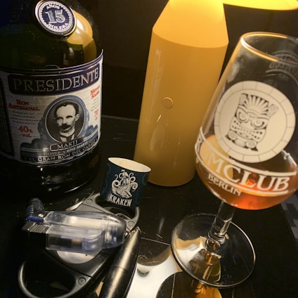 A bottle of Presidente rum is placed next to a yellow lamp and a glass with the words RUM CLUB Berlin printed on it. The glass contains a golden brown liquid. There is also a dark object that appears to be a bottle opener or cutter, and a small cup with a Kraken logo.