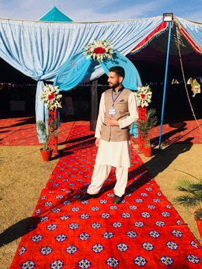 A person stands on a vibrant red carpet with intricate patterns. They are wearing traditional attire comprising a light-colored shalwar kameez and a brown waistcoat. The individual is in front of a decorated archway adorned with blue drapery and red and white floral arrangements. Potted plants are placed on either side of the carpet, leading into a tented area.
