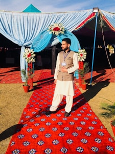 A person stands on a vibrant red carpet with intricate patterns. They are wearing traditional attire comprising a light-colored shalwar kameez and a brown waistcoat. The individual is in front of a decorated archway adorned with blue drapery and red and white floral arrangements. Potted plants are placed on either side of the carpet, leading into a tented area.