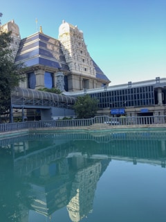 A grand temple structure with ornate, layered gopurams reflecting in a tranquil water body in the foreground. The architecture features traditional Indian styles with intricate carvings and a prominent use of blue and white colors. Trees and greenery partially surround the temple, adding a natural touch to the urban setting.
