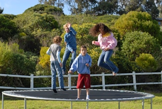 Four children are energetically bouncing on a trampoline surrounded by lush greenery and a white fence. The scene is full of movement, with one child in mid-air wearing a pink hoodie and jeans, and others dressed in casual clothing. The backdrop includes dense bushes and trees under a clear blue sky.