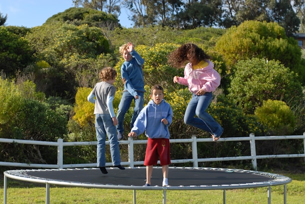 Four children are energetically bouncing on a trampoline surrounded by lush greenery and a white fence. The scene is full of movement, with one child in mid-air wearing a pink hoodie and jeans, and others dressed in casual clothing. The backdrop includes dense bushes and trees under a clear blue sky.