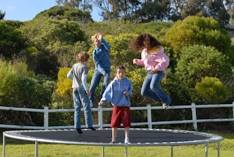 Four children are energetically bouncing on a trampoline surrounded by lush greenery and a white fence. The scene is full of movement, with one child in mid-air wearing a pink hoodie and jeans, and others dressed in casual clothing. The backdrop includes dense bushes and trees under a clear blue sky.