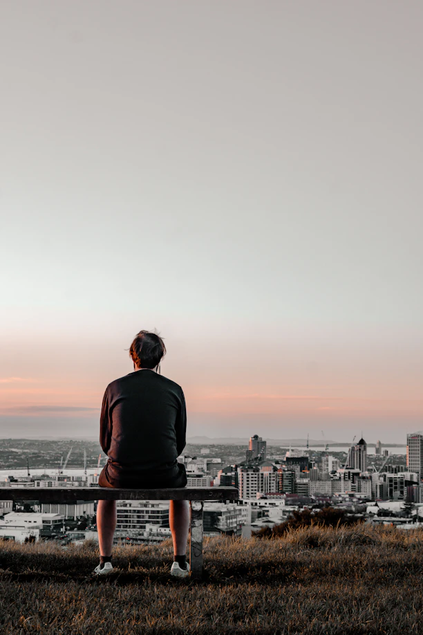 man in black long sleeve shirt sitting on brown wooden bench looking at city during daytime