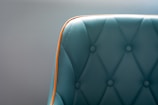 A teal upholstered chair with a quilted pattern and button detailing is highlighted by a bright orange trim. The focus is on the left section of the chair, with a soft gray background enhancing the elegant design.