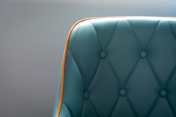 A teal upholstered chair with a quilted pattern and button detailing is highlighted by a bright orange trim. The focus is on the left section of the chair, with a soft gray background enhancing the elegant design.