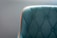 A teal upholstered chair with a quilted pattern and button detailing is highlighted by a bright orange trim. The focus is on the left section of the chair, with a soft gray background enhancing the elegant design.