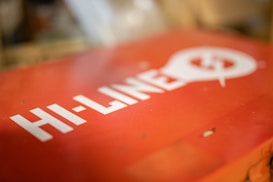 A red surface features bold white text that reads 'HI-LINE' alongside a stylized light bulb graphic with a power symbol. The background is blurred, drawing focus to the text and design.