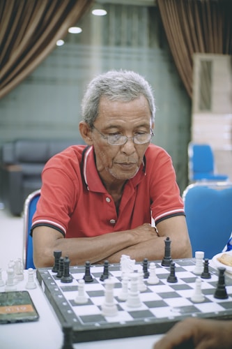 An elderly man wearing glasses and a red polo shirt focuses intently on a chess game. He is seated at a table with a chess board in front of him, displaying both black and white chess pieces. The background features blue chairs and a room with a softly lit ambience.