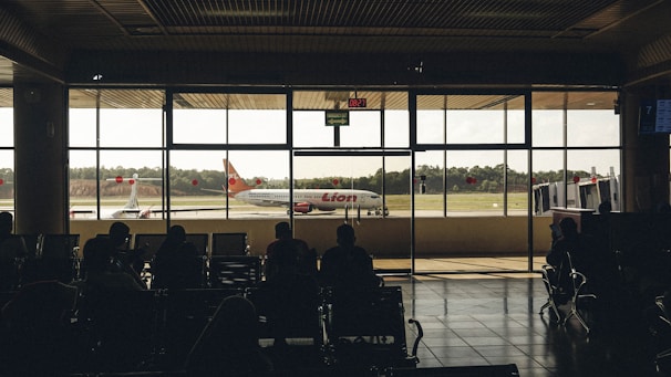 A dimly lit airport terminal with silhouettes of passengers seated and waiting, observing a Lion Air airplane positioned outside on the tarmac through large glass windows. The clock on the wall indicates the time as 08:21.