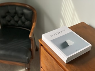 A stack of various books on a wooden table with a cozy reading nook.