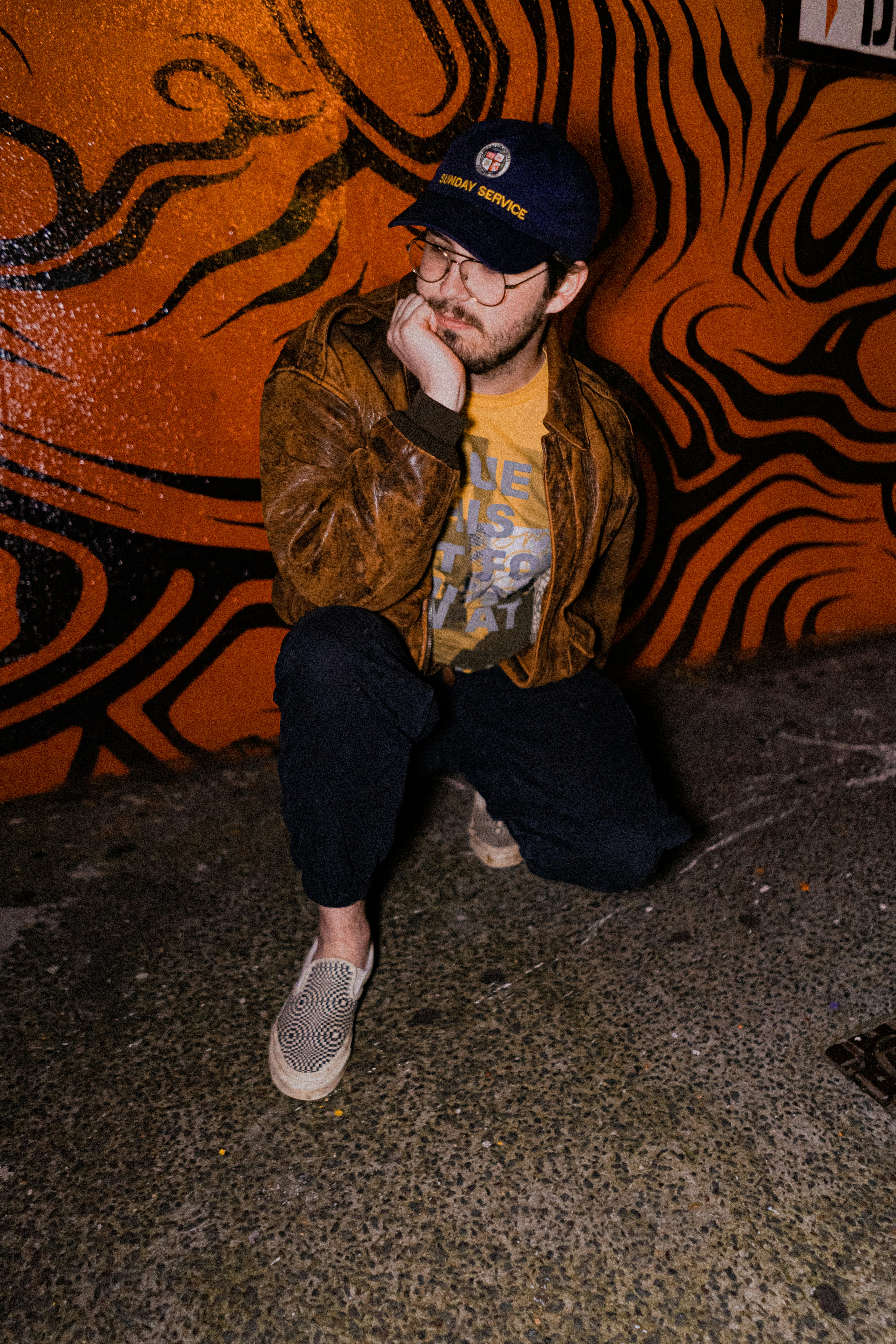 man in brown jacket and blue denim jeans sitting on brown concrete wall