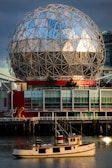 A geodesic dome structure with a reflective surface stands prominently against a backdrop of dark clouds and blue sky. Below the dome, there's a modern building with colorful panels featuring reds, blues, and greys. In the foreground, a classic wooden boat floats on calm water, reflecting the structure above.