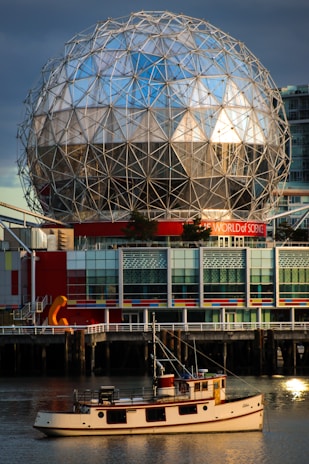 A geodesic dome structure with a reflective surface stands prominently against a backdrop of dark clouds and blue sky. Below the dome, there's a modern building with colorful panels featuring reds, blues, and greys. In the foreground, a classic wooden boat floats on calm water, reflecting the structure above.