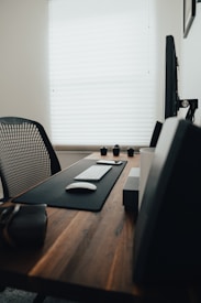 A modern office desk setup featuring a wooden table with a black mouse pad, keyboard, and mouse neatly arranged. There is a smartphone placed on the pad, and in the background, a monitor is visible. The room is softly lit through blinds, giving it a calm and organized appearance. A mesh chair is placed at the desk, and speakers and camera lenses are positioned along the back edge.