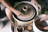 Hands covered in clay are shaping pottery on a spinning potter's wheel. Nearby, there are various tools and a sponge, indicating active involvement in the pottery-making process. The scene captures the focus and technique involved in crafting ceramics.