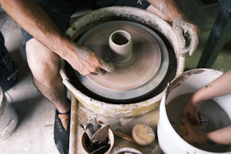 Hands covered in clay are shaping pottery on a spinning potter's wheel. Nearby, there are various tools and a sponge, indicating active involvement in the pottery-making process. The scene captures the focus and technique involved in crafting ceramics.