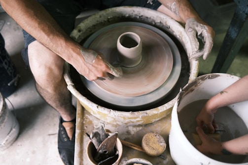 Hands covered in clay are shaping pottery on a spinning potter's wheel. Nearby, there are various tools and a sponge, indicating active involvement in the pottery-making process. The scene captures the focus and technique involved in crafting ceramics.