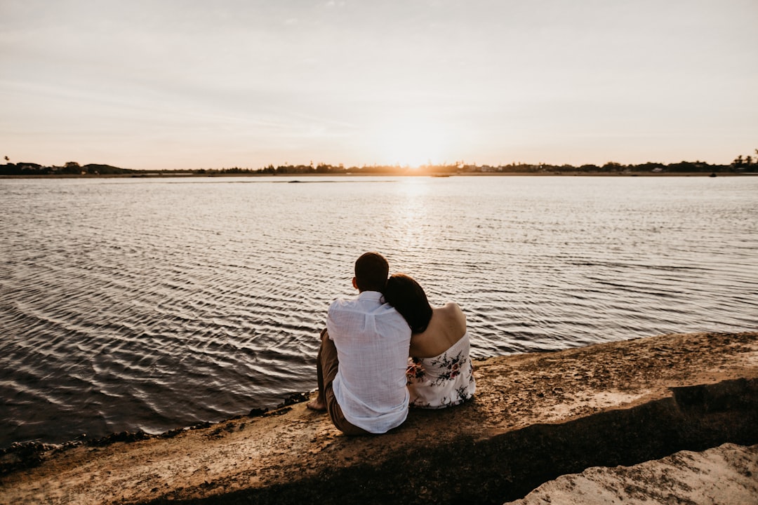 couple sitting on brown sand near body of water during daytime