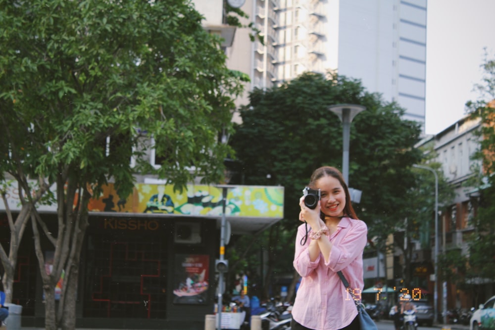 woman in pink coat holding black camera standing near green tree during daytime