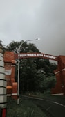 A brick archway with the words 'TUGU WISATA DESA RAHTAWU' spans across a road, surrounded by lush greenery and trees. A streetlight is positioned near the arch, and the sky appears overcast, creating a moody atmosphere.