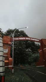 A brick archway with the words 'TUGU WISATA DESA RAHTAWU' spans across a road, surrounded by lush greenery and trees. A streetlight is positioned near the arch, and the sky appears overcast, creating a moody atmosphere.