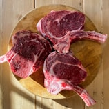 Three raw, bone-in ribeye steaks arranged on a round wooden cutting board, placed on a light wood table. The meat has a rich marbling and is partially in shadow, suggesting natural lighting.