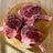 Three raw, bone-in ribeye steaks arranged on a round wooden cutting board, placed on a light wood table. The meat has a rich marbling and is partially in shadow, suggesting natural lighting.