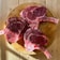 Three raw, bone-in ribeye steaks arranged on a round wooden cutting board, placed on a light wood table. The meat has a rich marbling and is partially in shadow, suggesting natural lighting.