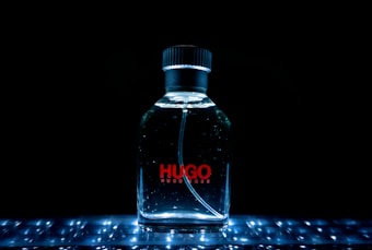 A clear glass bottle of Hugo Boss perfume, illuminated with bright lights against a dark background. The word 'HUGO' is prominently displayed in red on the bottle.