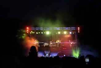 A live music concert at night featuring a band performing on a brightly lit stage. The stage is equipped with colorful lighting, including red, green, and blue hues, creating a vibrant atmosphere. The audience is silhouetted against the stage, with some spectators holding up phones to capture the moment.