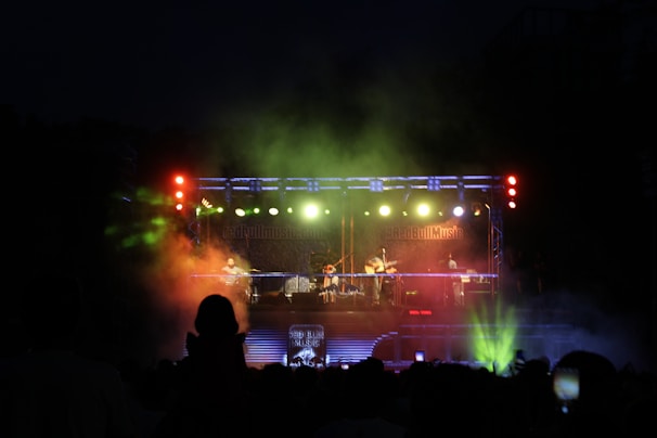 A live music concert at night featuring a band performing on a brightly lit stage. The stage is equipped with colorful lighting, including red, green, and blue hues, creating a vibrant atmosphere. The audience is silhouetted against the stage, with some spectators holding up phones to capture the moment.