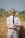 A person wearing a light-colored suit, dark sunglasses, and a black tie stands in front of a haystack in an outdoor setting. The individual is checking their watch and has a confident posture. The background shows a blurred natural landscape.