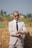 A person wearing a light-colored suit, dark sunglasses, and a black tie stands in front of a haystack in an outdoor setting. The individual is checking their watch and has a confident posture. The background shows a blurred natural landscape.