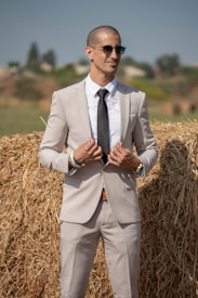 A confident man in a tan suit stands in front of a haystack, holding his suit jacket with both hands. He wears sunglasses and a black tie, and his demeanor suggests a relaxed yet stylish attitude. The background features blurred vegetation and a clear blue sky, contributing to a rustic and outdoor setting.