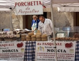 A market stall displays a variety of cheeses and other local food products. Two people are tending to the stall, with a man examining the goods and a woman standing beside him. The stall is covered with a checkered tablecloth, and several signs and banners advertise 'Porchetta da Pasqualino' and 'Salumi di Norcia'. There are stacks of cheese wheels and baskets of meats set on the table under a canopy.