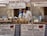 A market stall displays a variety of cheeses and other local food products. Two people are tending to the stall, with a man examining the goods and a woman standing beside him. The stall is covered with a checkered tablecloth, and several signs and banners advertise 'Porchetta da Pasqualino' and 'Salumi di Norcia'. There are stacks of cheese wheels and baskets of meats set on the table under a canopy.